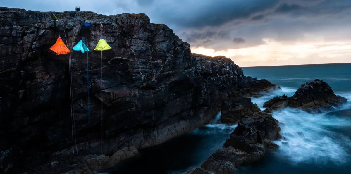 Colorful tents suspended on a rocky cliffside at twilight, overlooking a serene ocean.