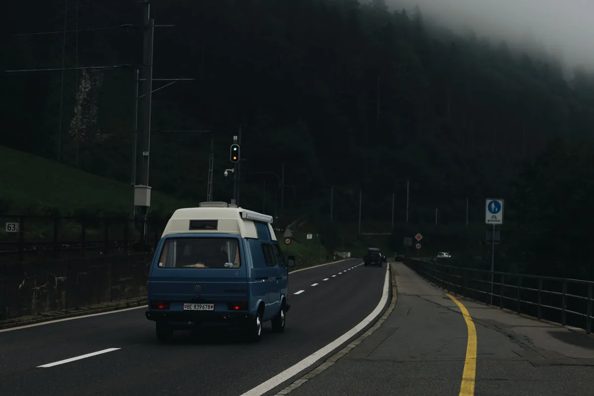 A blue camper van traveling on a misty road, surrounded by a foggy forest.