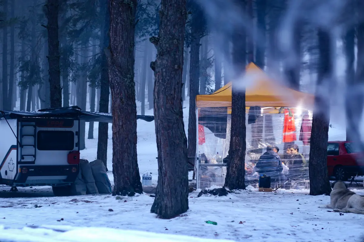 A cozy winter campsite with a tent and camper in a snowy forest setting.