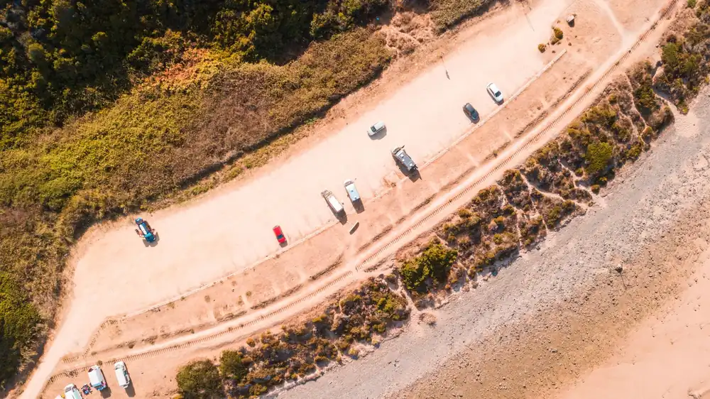 Aerial beachside parking view with cars lined along sandy shore and lush greenery.